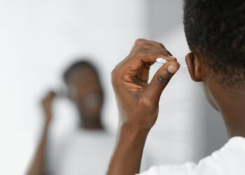 Black Man Cleaning Ears Using Cotton Swab Standing In Bathroom