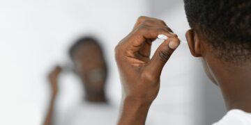 Black Man Cleaning Ears Using Cotton Swab Standing In Bathroom